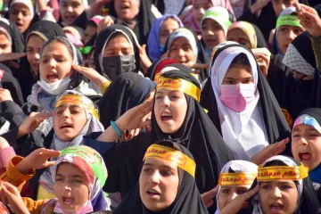 L'image montre un grand groupe de jeunes filles, portant des hijabs et des bandeaux avec des inscriptions en arabe. Elles semblent exprimer de l'enthousiasme, chantant ou criant, et leurs visages sont pleins d'énergie. L'arrière-plan indique un rassemblement, probablement lié à un événement spécial ou une manifestation. Les couleurs des vêtements et des bandeaux apportent une atmosphère vibrante à la scène.