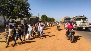 L'image montre une scène de rue animée dans une zone urbaine. Sur le côté gauche, on voit un arbre et des bâtiments, tandis que des gens marchent le long de la route en terre. Parmi eux, certains portent des sacs ou des charges sur la tête. Des véhicules, y compris des camions et des motos, circulent sur la route, créant une ambiance dynamique. L'éclairage semble être en plein jour, suggérant une activité quotidienne typique dans cette communauté.