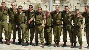 The image features a group of eleven soldiers standing closely together, wearing olive green military uniforms and boots. They appear to be in a celebratory mood, with smiles and casual poses. The background suggests a stone wall, possibly the Western Wall in Jerusalem, indicating a significant location. The soldiers carry some equipment, hinting at their military roles. Overall, the image conveys camaraderie and a sense of accomplishment among the group.