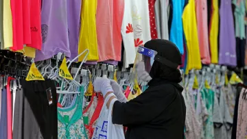 La imagen muestra a una persona en una tienda de ropa. Está usando una mascarilla y un protector facial. La persona está revisando prendas de vestir colgadas en perchas, que son de diversos colores y estilos. Se pueden ver camisetas y pantalones en el fondo, mientras que tiene una bolsa en la mano. La escena refleja un ambiente de compra de ropa, probablemente en un contexto donde se aplican medidas de seguridad relacionadas con la salud.