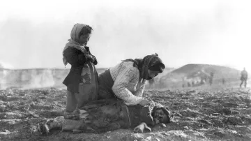 L'immagine mostra una scena drammatica e toccante. Una donna si trova inginocchiata su un terreno aperto, mentre si prende cura di un bambino disteso a terra, visibilmente in difficoltà. Un'altra bambina, in piedi accanto a loro, osserva con espressione seria. La scena è in un contesto desolato, con uno sfondo che suggerisce un ambiente di guerra o di sofferenza, evidenziando la vulnerabilità e il dolore delle persone coinvolte. I volti mostrano emozioni profonde che riflettono la situazione tragica in cui si trovano.