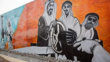 L'image représente une grande fresque murale. On y voit des figures en vêtements traditionnels arabes, probablement des hommes, qui semblent interagir avec des éléments symboliques liés à l'industrie pétrolière. L'arrière-plan est composé de couleurs vives, notamment des nuances de bleu et d'orange. Cette œuvre évoque des thèmes de culture, de tradition et de développement industriel, sur fond de paysages désertiques. La composition suggère un sens de la coopération et de l'engagement vers une vision commune.