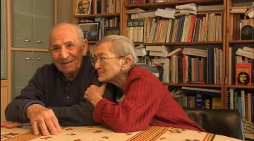 L'image montre un couple âgé assis à une table. L'homme a les cheveux gris et porte un pull sombre, souriant avec bienveillance. La femme, également âgée, a des cheveux très courts et porte un pull rouge. Elle est légèrement penchée vers lui, affichant une expression tendre et affectueuse. En arrière-plan, on peut apercevoir des étagères remplies de livres, créant une atmosphère chaleureuse et intellectuelle. La décoration présente un style simple et accueillant.