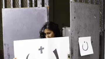 L'image montre une femme se tenant devant une porte grise. Elle tient une grande affiche présentant un symbole souriant, accompagnée d'un plus et d'un autre symbole. À côté, il y a une petite affiche accrochée à la porte. L'ensemble de la scène évoque une forme d'expression artistique, mélangeant des éléments de surprise et de réflexion.