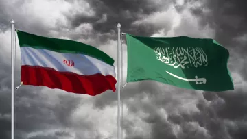 Nell'immagine sono visibili due bandiere: quella dell'Iran a sinistra e quella dell'Arabia Saudita a destra. Entrambe le bandiere sono sovrapposte a uno sfondo scuro e nuvoloso, creando un contrasto suggestivo. La bandiera iraniana è composta da bande orizzontali di verde, bianco e rosso, con lo stemma nazionale al centro. La bandiera saudita è verde, con la scritta in arabo e una spada bianca. L'insieme trasmette una sensazione di tensione e rivalità tra i due paesi.