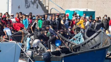 La imagen muestra un grupo de personas en un puerto o zona costera, donde hay varias embarcaciones. Muchas de las personas parecen estar en una o varias pequeñas embarcaciones de madera, mientras que otras están en la orilla o cerca de las embarcaciones. Se observa una multitud, con algunas personas vestidas en ropa casual, y parece que están en un momento de actividad o espera. Al fondo, hay estructuras de concreto y un baño portátil. La escena sugiere un ambiente de migración o búsqueda de oportunidades.