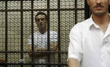 Deux hommes se tiennent derrière des barreaux, l'un visible et l'autre en arrière-plan.