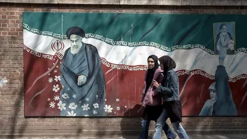 L'image montre un mur décoré d'une peinture murale qui représente un personnage historique en relation avec l'Iran, entouré de motifs floraux et de couleurs représentant le drapeau iranien. Au premier plan, deux femmes marchent le long du mur, vêtues de vêtements modernes et de foulards, illustrant un contraste entre le patrimoine culturel et la vie contemporaine. L'ambiance de la scène évoque un mélange de tradition et de modernité dans le cadre urbain iranien.