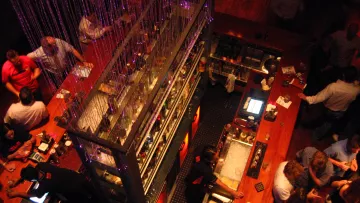 L'image montre une vue aérienne d'un bar animé. On peut voir une grande table de bar en bois clair, avec de nombreuses personnes stationnées autour, se parlant et déguster des boissons. Au dessus, il y a des chaînes de lumières, créant une ambiance festive. Les étagères à bar sont bien garnies de bouteilles colorées, et on distingue des serveurs s'affairant derrière le comptoir. L'atmosphère semble conviviale et dynamique, avec une décoration moderne.