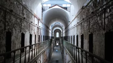 L'image montre l'intérieur d'une ancienne prison, avec un long couloir central bordé de cellules. Les murs sont décolorés et décrépis, témoignant de l'âge de l'établissement. Le plafond est haut, avec de grandes arches et une lumière qui pénètre par des lucarnes au-dessus. Les balustrades en fer forgé entourent les niveaux supérieurs, ajoutant à la structure vétuste. L'ambiance générale suggère une atmosphère d'abandon et d'histoire.