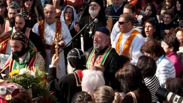 L'image représente un groupe de personnes rassemblées lors d'une cérémonie religieuse. Au centre, des figures cléricales en vêtements liturgiques colorés chantent et prient, entourées de fidèles. On aperçoit des chants, des bougies et des fleurs décoratives, ce qui suggère un événement festif ou sacré. L'ambiance est chaleureuse et communautaire, avec une multitude de visages exprimant la dévotion.