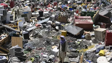 L'image montre un vaste amoncellement de déchets et de débris dans un espace encombré. On y voit des objets divers tels que des appareils électroménagers, des câbles, des pneus, et d'autres débris. Il y a aussi une personne qui marche au milieu de ce désordre, donnant une idée de l'échelle des détritus présents. L'environnement semble désordonné et négligé, illustrant une scène de récupération ou de désindustrialisation.