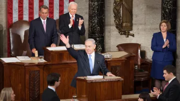 Cette image montre une scène dans une salle législative. Un homme se tient au pupitre, levant la main en signe de salut ou d'allocution. Derrière lui, plusieurs personnes, dont un homme plus âgé et une femme, l'écoutent attentivement. On peut également voir un drapeau américain en arrière-plan, ainsi que le mobilier typique d'une assemblée. L'atmosphère semble formelle et solennelle, suggérant un moment important, potentiellement un discours ou une présentation officielle.