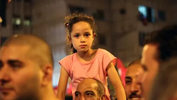 L'image montre une petite fille avec des cheveux bouclés, assise sur les épaules d'un homme dans une foule. Elle porte un t-shirt rose et a une expression sérieuse. Autour d'elle, on peut voir des visages d'adultes, certains souriant et d'autres concentrés, ce qui suggère une ambiance dynamique, peut-être lors d'un événement ou d'une célébration. L'arrière-plan est flou, mais on peut distinguer des lumières et des drapeaux, ajoutant à l'atmosphère animée de la scène.