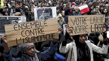 L'image montre une grande manifestation avec une foule dense de personnes rassemblées. Au premier plan, deux participantes tiennent des pancartes en bois sur lesquelles on peut lire des messages exprimant des revendications. L'une des pancartes dit "ENSEMBLE CONTRE L'ISLAMOPHOBIE" et l'autre "VOTRE LAÏCITÉ, VOTRE LIBERTÉ". Le contexte semble être une protestation pour défendre des droits liés à la laïcité et à la lutte contre la discrimination. Les gens portent des banderoles et des drapeaux, et l'atmosphère paraît déterminée et engagée.