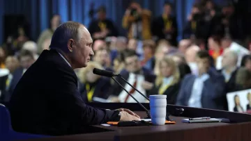 L'image montre un homme assis à une table de conférence, parlant peut-être lors d'une conférence de presse ou d'un discours. Il semble concentré et fait face à un public, qui est partiellement visible en arrière-plan. On aperçoit des journalistes prenant des notes ou photographiant. Des micros sont placés devant lui, et une tasse blanche est posée sur la table. L'atmosphère paraît sérieuse et attentive.