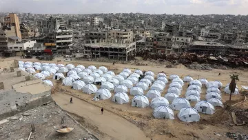 L'image montre une vaste zone de tentes blanches installées sur un terrain aride, entourée de bâtiments en ruines. Les tentes semblent former un camp, probablement pour des personnes déplacées ou des réfugiés. En arrière-plan, on peut voir des structures détruites, ce qui indique un environnement de conflit ou de catastrophe. Le ciel est nuageux, et l'atmosphère générale évoque la désolation et la nécessité d'abri.