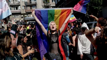 L'image montre une scène dynamique d'une marche ou d'une manifestation célébrant la fierté. Au centre, une personne tient une grande drapeau arc-en-ciel, symbole de la communauté LGBTQ+. Elle exprime sa joie ou son enthousiasme, visiblement impliquée dans l'événement. Autour d'elle, d'autres participants portent également des drapeaux et affichent des expressions de soutien et de célébration. L'environnement urbain en arrière-plan suggère que l'événement se déroule dans une ville. Les gens semblent heureux et engagés dans une atmosphère festive et inclusive.