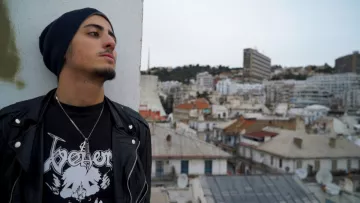 L'image montre un jeune homme qui se tient sur un toit, regardant au loin. Il porte un bonnet et une veste en cuir noire, ainsi qu'un t-shirt avec un design graphique. En arrière-plan, on aperçoit une ville avec des bâtiments de différentes tailles et architectures, et des toits de maisons. Le ciel est légèrement nuageux, ce qui donne une ambiance calme et contemplative à la scène.