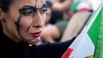 En la imagen se puede ver a una mujer con un maquillaje artístico que representa un mapa de México en su rostro, con un trazo destacado en negro y una lágrima roja que corre por su mejilla. Ella sostiene una bandera de México, que se encuentra en primer plano, mientras que en el fondo se vislumbran otras personas y un ambiente de manifestación. Su expresión es seria y reflexiva, lo que sugiere un fuerte mensaje emocional o político.