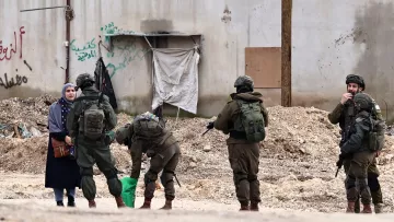 L'image montre une femme vêtue d'une robe sombre, se tenant à côté d'une rue. Devant elle, plusieurs soldats en uniforme sont regroupés, certains sont en train de fouiller un sac, tandis que d'autres observent la femme. En arrière-plan, on peut voir un mur avec des graffitis et des débris sur le sol.