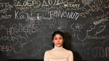 L'image montre une jeune femme debout devant un mur noirci couvert d'inscriptions multilingues. Elle porte un pull beige et a les cheveux longs et noirs. Le mur est recouvert de mots de différentes langues, créant un effet vibrant et accueillant. La femme semble sereine et posée, ajoutant une dimension humaine à ce mélange culturel représenté par les écritures.