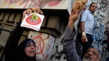 The image depicts a protest scene where individuals are holding up flatbreads and a sign. The sign features a dollar symbol with a prohibition sign over it, conveying a message against inflation or economic hardship, likely related to rising food prices. In the background, there are graffiti-covered walls, indicating a tense atmosphere. The protesters express their discontent, while another person appears in the background observing the scene.