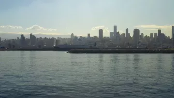 L'image montre un paysage urbain d'une ville moderne en bord de mer. On peut voir des gratte-ciels s'élevant le long de la côte, reflétant la lumière du soleil sur l'eau calme de la mer. Au premier plan, il y a un port ou une jetée, et l'arrière-plan est rempli de bâtiments qui se dressent majestueusement. Le ciel est légèrement nuageux, ce qui crée une ambiance douce et mélancolique. L'ensemble donne une impression de tranquillité et de modernité.