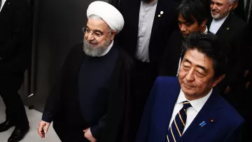 L'image montre deux hommes marchant côte à côte dans un environnement formel. L'un d'eux porte une tenue sombre avec une coiffure distinctive et un turban blanc, tandis que l'autre est habillé de manière formelle en costume. Ils semblent être entourés de plusieurs autres personnes habillées aussi de manière professionnelle. L'atmosphère dégage un air de réunion ou d'événement officiel.