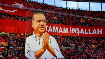 La imagen muestra un gran despliegue en un estadio, donde se aprecia una multitud de personas sosteniendo banderas rojas. En el centro, destaca un enorme cartel que presenta la figura de una persona aplaudiendo, acompañado de la frase "Tamam İnşallah", que se traduce como "Está bien, si Dios quiere." El ambiente parece ser festivo y de apoyo a esta figura, probablemente un líder político, en un evento significativo.