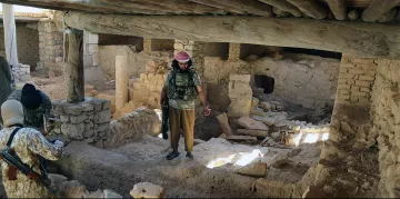 L'image montre un intérieur d'un bâtiment qui semble ancien et en ruine. On peut voir des murs en pierre et des débris éparpillés sur le sol. Un homme, habillé en vêtement militaire avec un bandeau, se tient au centre de la pièce, tandis que deux autres personnes, cachées en partie, l'observent. La lumière entre par des ouvertures, créant un contraste entre les ombres et les zones illuminées dans cet environnement désolé.