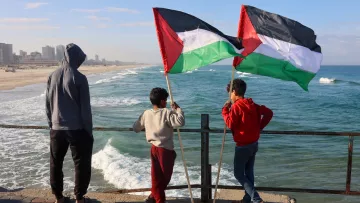 Nell'immagine, tre ragazzi sono in piedi su un molo che si affaccia su una spiaggia e sul mare. Due di loro tengono in alto delle bandiere palestinesi, che sono di colore nero, bianco, verde e rosso. Il ragazzo più piccolo, in una felpa grigia, sembra osservare attentamente l'orizzonte. La scena è illuminata da una luce naturale, con il cielo blu e alcune nuvole sparse. Le onde del mare si infrangono dolcemente sulla riva, creando un'atmosfera tranquilla. Gli altri due ragazzi indossano abbigliamento casual e sembrano condividere un momento di riflessione insieme.