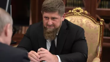 L'image montre un homme avec une barbe, assis à une table dans un cadre formel. Il est habillé en costume noir et semble concentré sur la discussion. En arrière-plan, on peut apercevoir des meubles élégants et une ambiance sophistiquée. L'éclairage est doux, ce qui contribue à l'atmosphère sérieuse de la scène.