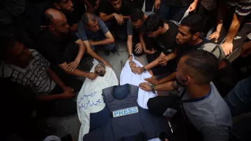 Des hommes entourent un corps recouvert, en deuil, avec une veste de presse à côté.