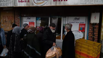 L'image montre une scène animée devant une boulangerie à Istanbul. On aperçoit une file de personnes, majoritairement des personnes âgées, qui attendent pour acheter du pain. Certains portent des sacs en plastique contenant des baguettes. Les gens semblent discuter entre eux, et la boulangerie affiche des panneaux avec des indications sur les produits. L'atmosphère est conviviale et caractéristique des marchés de quartier dans la ville.