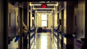 L'image montre un couloir d'hôpital, avec des murs clairs et un sol brillant qui reflète la lumière. Au loin, on aperçoit une fenêtre et une chaise. Les portes sur les côtés sont fermées, et une signalisation de sortie est visible au-dessus. L'ambiance est plutôt calme et légèrement sombre, accentuée par les lumières artificielles.