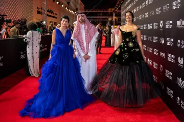 L'image montre trois personnes posant sur un tapis rouge lors d'un événement. À gauche, une femme porte une robe de soirée bleue élégante avec des volants. Au centre, un homme habillé en tenue traditionnelle, comprenant un thobe et un chapeau saoudien. À droite, une femme porte une robe noire ornée de motifs floraux, ajoutant une touche sophistiquée à son look. L'arrière-plan montre un décor stylisé, typique des événements glamour.