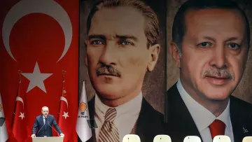 L'immagine ritrae un evento politico in Turchia. Sullo sfondo, sono presenti due grandi ritratti: uno di Mustafa Kemal Atatürk, fondatore della Repubblica turca, e l'altro di Recep Tayyip Erdoğan, attuale presidente. Al centro, un oratore sembra parlare al pubblico, con bandiere turche e simboli del partito sul palco. Lo sfondo con i due leader storici suggerisce un legame tra il passato e il presente politico del paese.