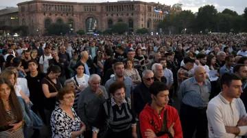 L'image montre une grande foule de personnes rassemblées dans un espace public, probablement pour un événement ou une manifestation. Les gens semblent concentrés et attentifs, certains regardant vers l'avant. L'arrière-plan présente des bâtiments, suggérant un milieu urbain, et le ciel est légèrement nuageux, indiquant que le jour commence à tomber. L'atmosphère semble à la fois solennelle et engagée.