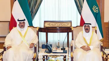 The image shows two men seated in a formal setting, likely in a government or diplomatic office. Both are wearing traditional attire, including white thobes and head coverings. They are positioned next to each other, with an ornate decorative background featuring curtains and a rich interior design. On the table in front of them, there are flowers and a few items, including a telephone. Behind them, a plaque with Arabic script can be seen, indicating the formal nature of the meeting.