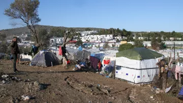 L'image montre une scène d'un campement de migrants. On peut voir des tentes montées sur un terrain plutôt nu et chaotique, avec des déchets éparpillés autour. À l'arrière-plan, des bâtiments sont visibles, suggérant qu'il y a une communauté près de ce camp. Le ciel est clair et ensoleillé, et il y a quelques arbres et collines dans le paysage. Une personne se tient debout dans la scène, semblant observer les environs. L'ensemble dégage une impression de précarité et de lutte.