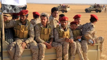 L'image montre un groupe de soldats assis à l'arrière d'un véhicule militaire dans un environnement désertique. Ils portent des uniformes camouflés et des casques ou des bérets rouges. À l'arrière-plan, on aperçoit d'autres véhicules militaires. Le paysage est aride et sablonneux, typique des zones désertiques. Les soldats semblent concentrés et disposés à exécuter leur mission.