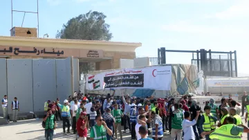 L'image montre une scène de manifestation à un point de passage, probablement en rapport avec des événements politiques. On peut voir des gens rassemblés, certains portant des gilets fluorescents, et une grande banderole avec un message en arabe. Des drapeaux, dont celui de l'Égypte, flottent à proximité. Le cadre semble être une zone de contrôle ou un poste de frontière, et l'ambiance est animée avec des personnes qui discutent ou manifestent.