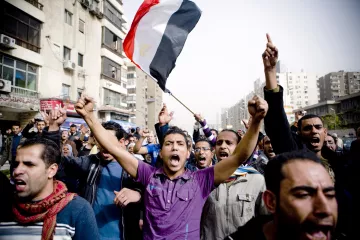 الصورة تظهر مجموعة من الناس في مظاهرة حاشدة. في المقدمة، يوجد رجل يحمل علم مصر ويبدو أنه يرفع صوته عالياً تعبيراً عن الحماس أو المطالب. خلفه يظهر حشد كبير من المتظاهرين، بعضهم يرفعون أيديهم ويهتفون. تعكس الصورة أجواء من النشاط والمشاركة الشعبية، مع مباني المدينة في الخلفية تحت سماء واضحة.
