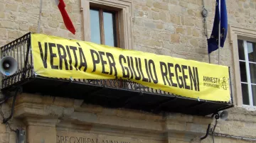 L'immagine mostra un grande striscione giallo appeso a un balcone di un edificio. Sullos striscione è scritto "VERITÀ PER GIULIO REGENI", accompagnato dal logo di Amnesty International. Nel contesto, si possono notare delle bandiere, probabilmente italiane, che sventolano. L'atmosfera suggerisce una manifestazione di sostegno e richiesta di giustizia per Giulio Regeni, un caso di rilevanza pubblica.
