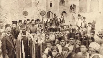 L'image montre un groupe de personnes rassemblées dans une ruelle étroite, probablement d'une ville historique. Au premier plan, on peut voir des hommes en habits traditionnels, certains portant des chapeaux typiques. Un homme central semble être un dignitaire religieux, accompagné d'une procession. En arrière-plan, des enfants et d'autres adultes regardent vers l'objectif, créant une atmosphère de communauté et de partage. Les bâtiments en pierre autour d'eux suggèrent un contexte ancien.