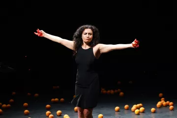 L'image montre une femme debout sur scène, les bras étendus de chaque côté. Elle porte une robe noire et exprime une émotion intense. Des oranges sont dispersées autour d'elle sur le sol, ajoutant une dimension visuelle frappante à la scène. Des éclaboussures de liquide rouge semblent provenir de ses mains, ce qui accentue le drame et la tension de l'instant. L'éclairage de la scène est sombre, mettant ainsi en valeur la silhouette de la femme et les oranges.