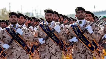 The image shows a group of uniformed soldiers in military attire. They are wearing camouflage patterns and black berets, and some are adorned with blue and gold sashes. The soldiers are standing in formation, holding rifles and displaying a serious demeanor. The background appears to have a clear sky and possibly some military infrastructure, emphasizing a display of military presence or a ceremony.