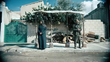 L'image montre une scène dans une rue, où deux personnes se tiennent sous un abri. À gauche, une femme est vêtue d'une longue robe sombre et porte un foulard, tandis qu'à droite, un homme, habillé de manière militaire, se tient debout, les mains derrière le dos. En arrière-plan, on voit un mur en briques avec des débris et des plantes, notamment un cactus. L'environnement a une ambiance un peu désolée et montre des signes d'abandon. Le ciel est partiellement nuageux, ajoutant une certaine atmosphère à la scène.