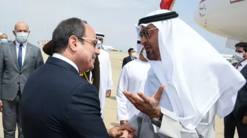 L'image montre une rencontre entre deux dirigeants. Ils semblent être en conversation amicale, avec des gestes visibles pour accompagner leurs paroles. L'un des deux est vêtu d'un costume formel, tandis que l'autre porte des vêtements traditionnels arabes, incluant un keffieh. En arrière-plan, on peut apercevoir des membres du personnel et des avions, suggérant que la scène se déroule dans un aéroport. L'atmosphère paraît conviviale et respectueuse.